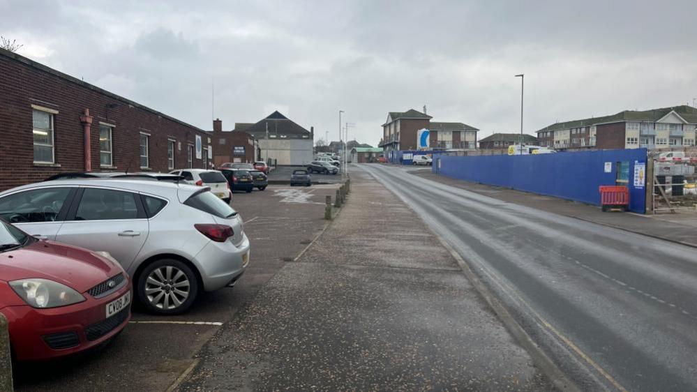 The Conge, showing Downey's printing works and a blue hoarding on the south side of the street where development to housing is set to go ahead subject to planning permission. Cars are parked on the left of the image, and the road runs through the centre/right of the picture.