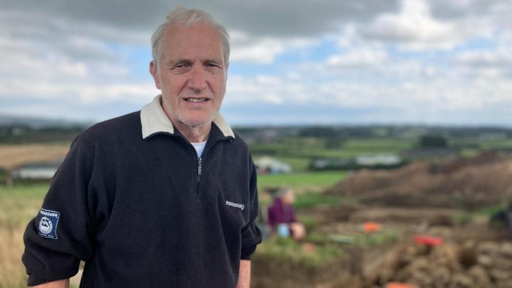 An older man wearing a black jumper stands in front of an archaeological site