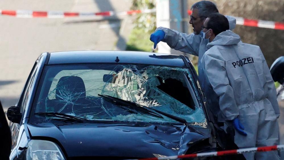 Two policemen in white hazmat suits inspect a black car with a broken windshield and heavily dented hood