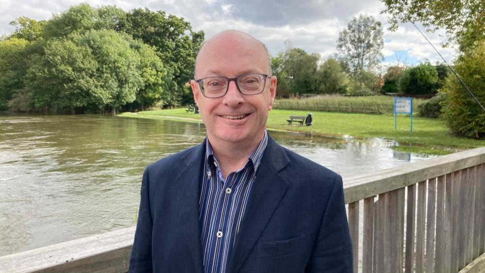 Simon Clewlow, standing on a bridge, over water, with greenery in the back ground. He is looking at the camera and smiling and wearing a blue and white striped shirt and blue jacket.