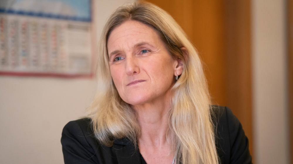 A woman with long blonde hair looks away from the camera. She is wearing a black blazer and sitting in an office.