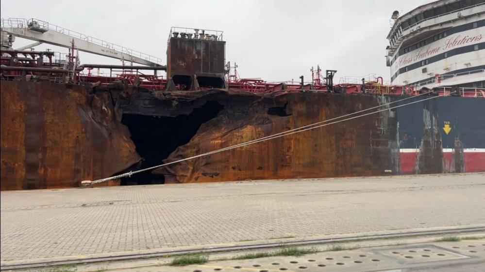 The significant hole in the side of the Stena Immaculate, at rest on the North terminal of Great Yarmouth's outer harbour. The hull is rusting and fire damaged following a collision in March off the Humber Estuary. The ship superstructure and bridge is visible to the right of the image. Quayside hardstanding is visible in the foreground.
