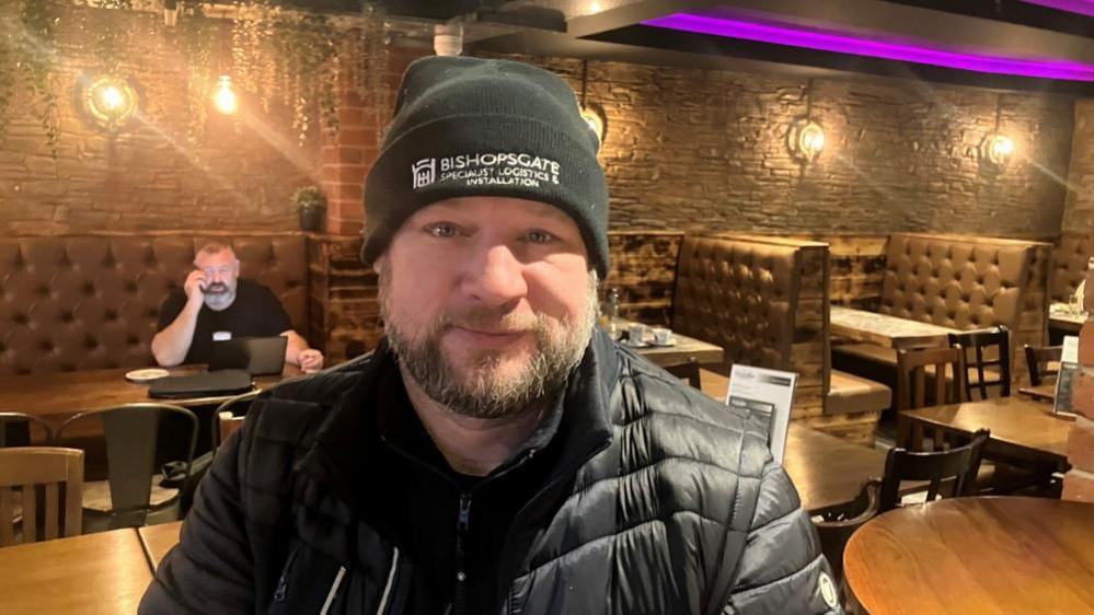 A man in his eary 40s sits in a cafe. He is wearing a black puffer jacket and a beanie. 