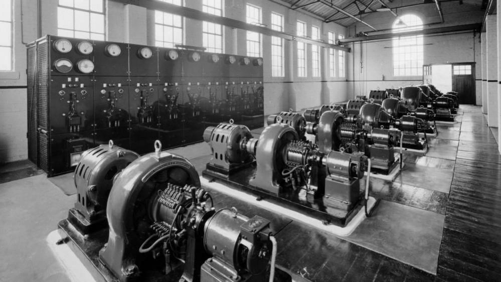 A line of nine spinning generator machines made of metal. They are alongside tall black equipment racks with dials at the top. There are windows along the wall to the left and a large cathedral window at the end of the building in this black and white picture.