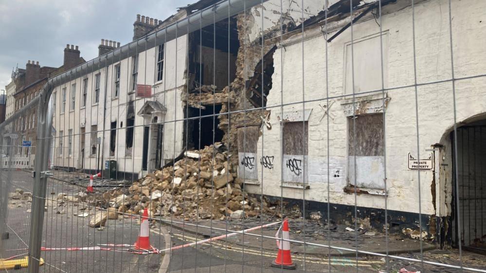 Two two-storey white houses side by side. There is a gap in the front facade in which the base of an upper floor can be seen. There is a large pile of rubble including bricks in front of the gap, spilling onto the pavement. The building is fenced off.