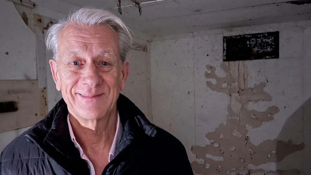 Trevor Blythe, in a black jacket and pink shirt, standing in the bunker, and the ventilation shaft to the left of the image.