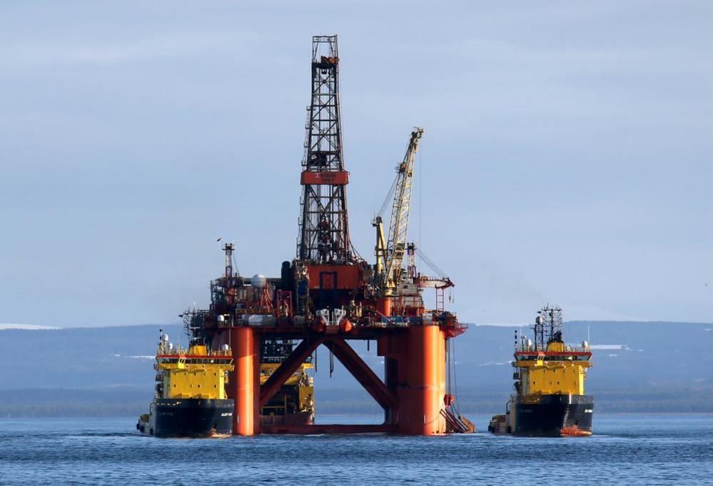 Oil platform with rigging, with yellow boats on either side. The platform has an orange-coloured base which extends into the blue sea. There is land in the distance and high cloud in the sky.