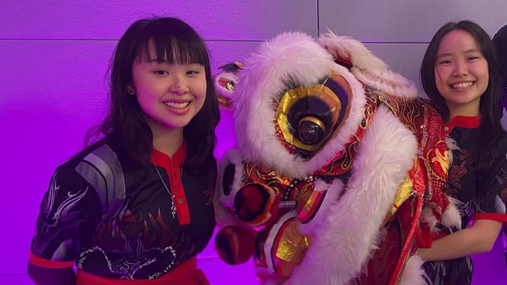 Two Bristol lion dance troupe members wearing black and red outfits, holding a white fury lion head mascot against a purple background.