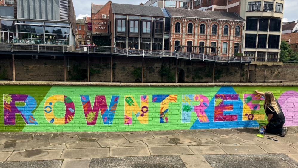 A view of a vibrant mural, painted along a low brick wall along a riverside path. The mural depicts the word 'Rowntree' painted in bright multicolours against predominately lime-green background. Each letter also contains leaf and flower motifs within it. On the opposite side of the river, a metal walkway and several buildings housing bars can be seen. In the corner, a woman with long blonde hair and wearing black paints onto the mural.