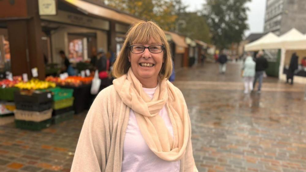 Wendy Randall with medium-length brown hair and glasses, smiling and wearing a yellow wrap-around scarf and cardigan with a fruit market stall in the background