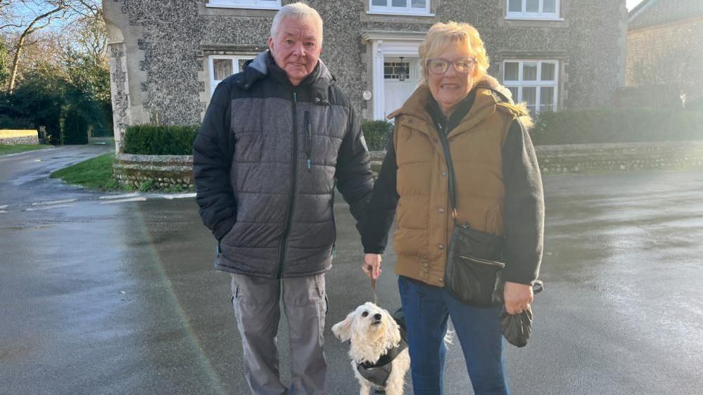 Dennis and Linda Hammond, standing in the centre of a village, holding a small dog, who is in the middle of them. Dennis is wearing a dark coat, with grey trousers, Linda, a dark top with an orange gilet over the top. She has a black handbag across her, and is holding a black dog waste bag in her left hand. They are both looking straight at the camera, with a flint stone building behind them and a road. 