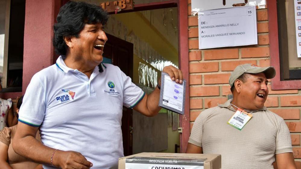 Morales laughs as he holds up his ballot paper, about to put it into the ballot box. An election official is standing behind him, also laughing - they're both looking off to the side at something.