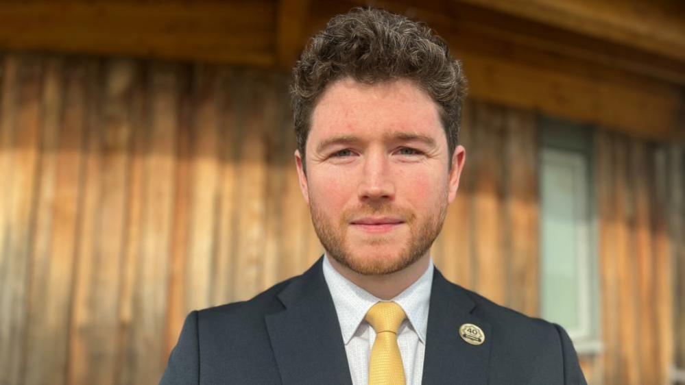 Ross Barton, looking at the camera, wearing a dark suit, with a yellow tie, he has dark hair and a beard 