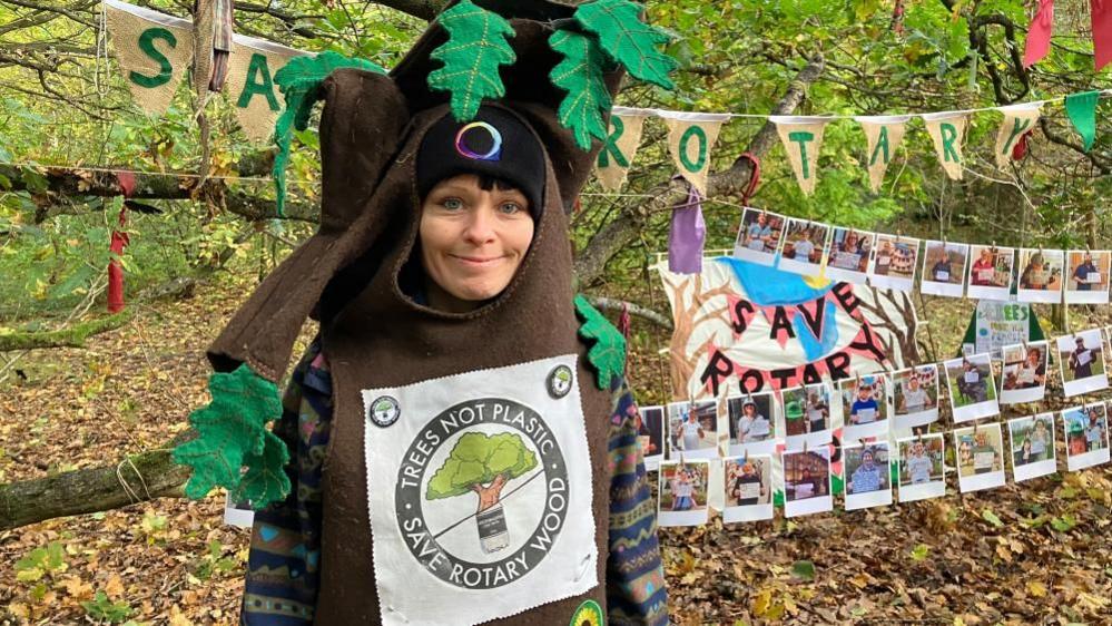She wears a tree costume; brown trunk body with felt leaves. She stands in front of a campaign display in the woodland, including banners and bunting spelling out 'save Rotary Wood', as well as photography pinned to washing line-type string tied across branches.