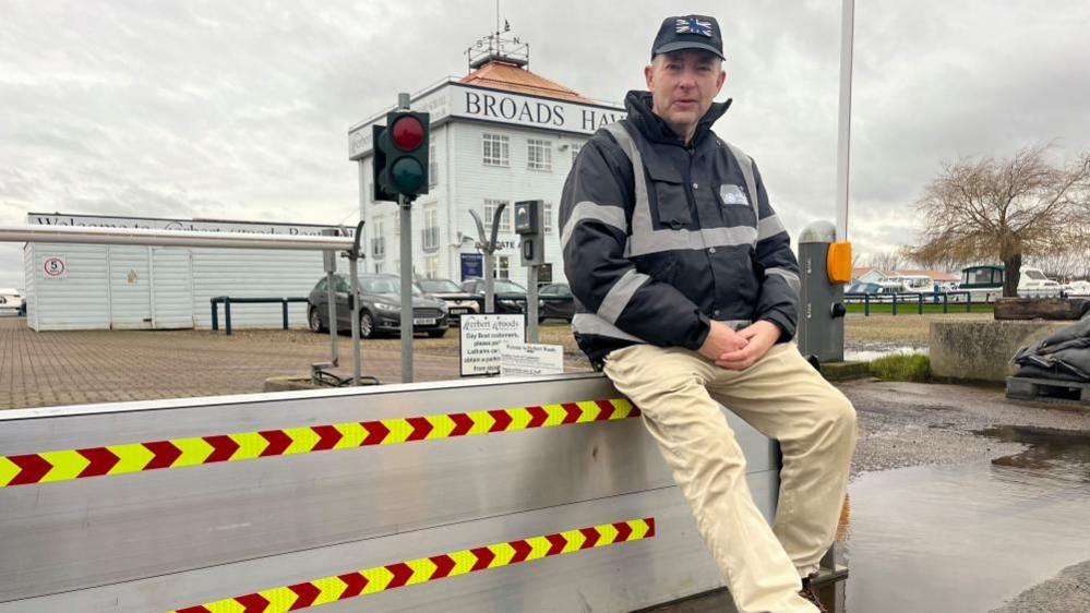 Paul Rice, sitting on the flood barrier at Potter Heigham. He is wearing a black waterproof jacket with high visibility markings and cream coloured trousers, and a blue baseball cap. Behind him is the Broads Haven building.s