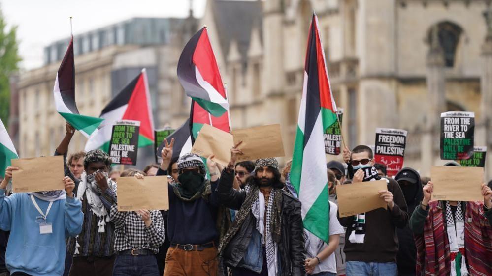 Protest in Cambridge: protesters, with some wearing scarves and dark glasses, hold up signs and red, green and white flags.
