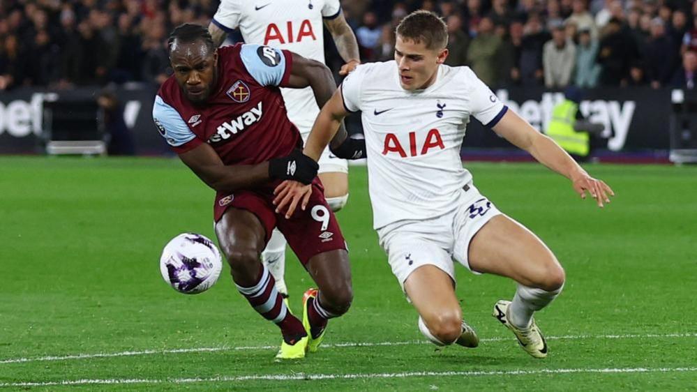 Player in red West Ham strip and player in white Tottenham strip attempt to get the ball, which is airborne above the football pitch. Spectators are visible behind.