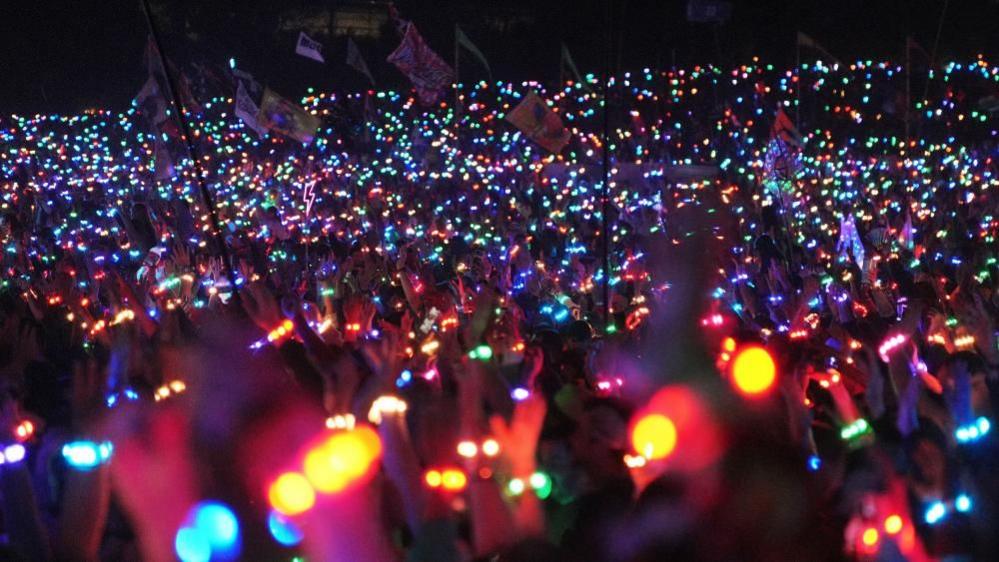 A crowd of music fans in the dark are raising their hands in the air, all wearing bracelets that are glowing different colours. Some are waving flags, which are visible in the background.