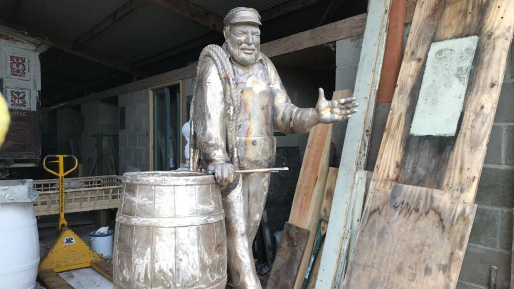 A bare metal sculpture of artist Ernie Childs, featuring him wearing his trademark cap, and holding his left hand out. He has his right hand on a herring barrel. The picture is taken within a shed, with board and timber stacked up against the wall on the right of the image, and a yellow pallet truck to the left of the image, with other detritus in the background.