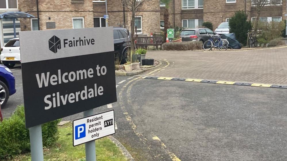 A white and grey sign saying "Welcome to Silverdale" in front of brown-bricked buildings and cars. A smaller sign says "Resident permit holders only".