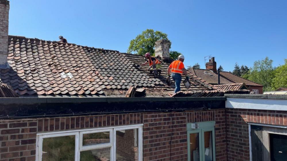 Two workers on the rear roof of the Decoy Tavern, dressed in high visibility clothing, and a third person whose head and shoulders are visible above the ridge line, on the front side of the building. The tiles are being removed. Windows and doors are visible in the image, and a chimney stack.