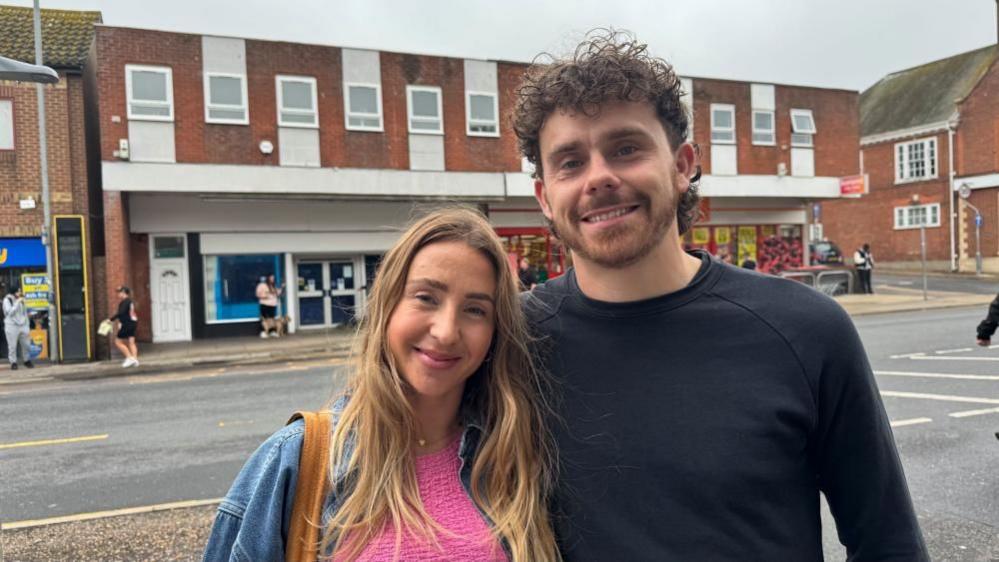 Lily Duncan is wearing a denim jacket and pink jersey, and has light brown bag straps over her right shoulder. She has long strawberry blonde hair and is smiling. She is standing close to her husband Jordan, who is wearing a black long sleeve T-shirt. He has long dark hair and a dark/ginger full beard.