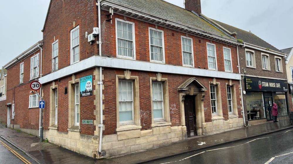 The former Barclays on Gorleston High Street is a 1920s building with red brickwork and stone mullions and foundation work. It has a Georgian-style entrance and a white fascia which once bore the Barclays sign. Above that white band is a first floor of sash windows, leading to a slate roof