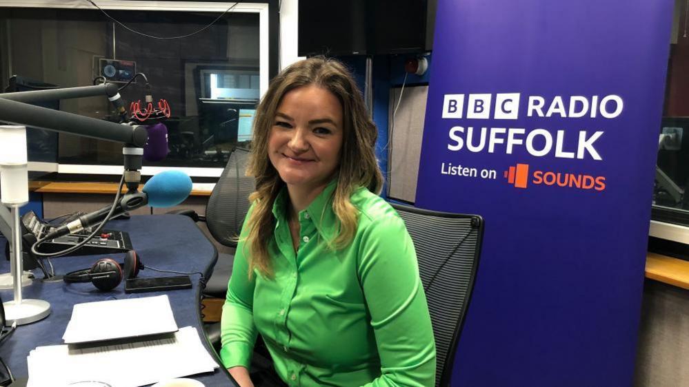 Jenny Riddell-Carpenter is sitting in the BBC Radio Suffolk studios. She has long blonde hair and is smiling at the camera She is wearing a bright green shirt. 