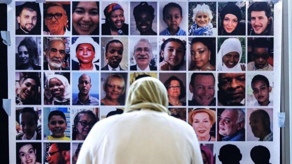 A woman stands with her back to the camera looking at a wall of photos of the people killed in the Grenfell fire