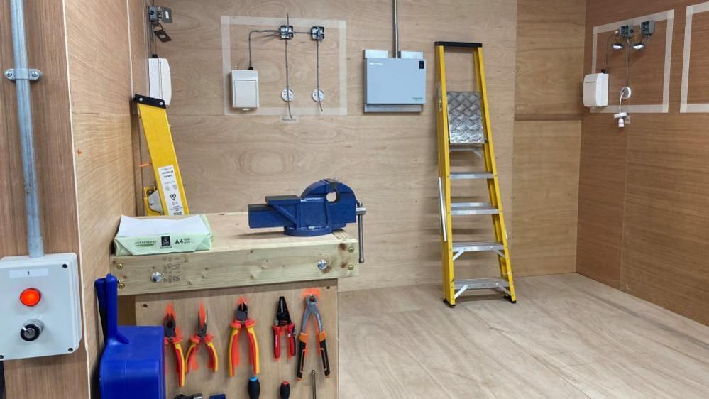 A view of one of the cubicles students learn in, showing a step ladder, various pliers hanging up, and sockets on the wall