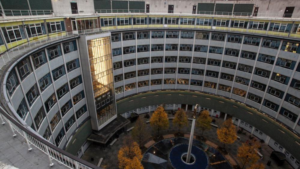 A U-shaped building with lots of dark windows and cladding, there are trees in the middle section and a large rectangular lift.