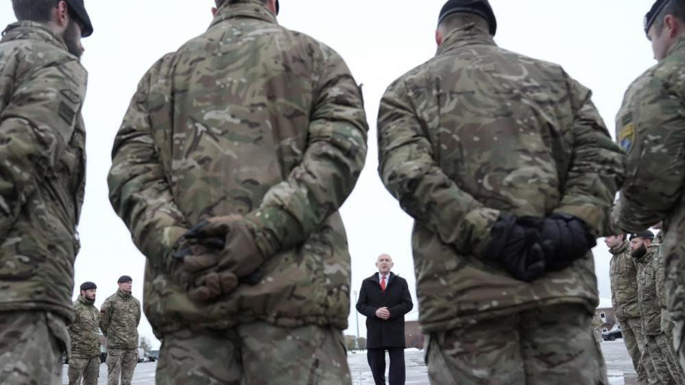 Soldiers stand in a circle listening to a man wearing a tie - the UK's defence secretary John Healey