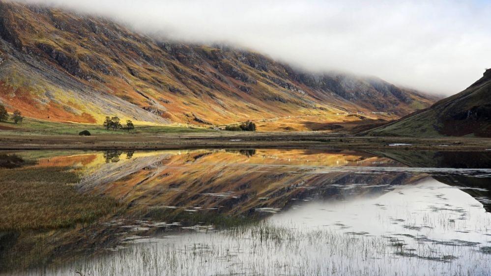 Golden hills reflected in the water of a loch with a cloudy sky