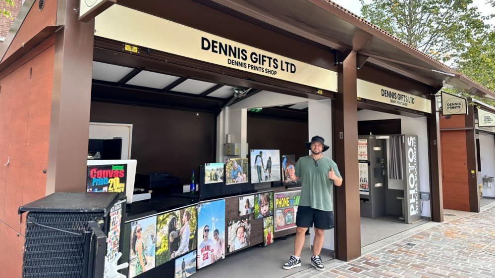 Man in green hat and T-shirt standing with thumbs up in front of a wood-coloured stall selling paintings. Various pieces of artwork are on display.