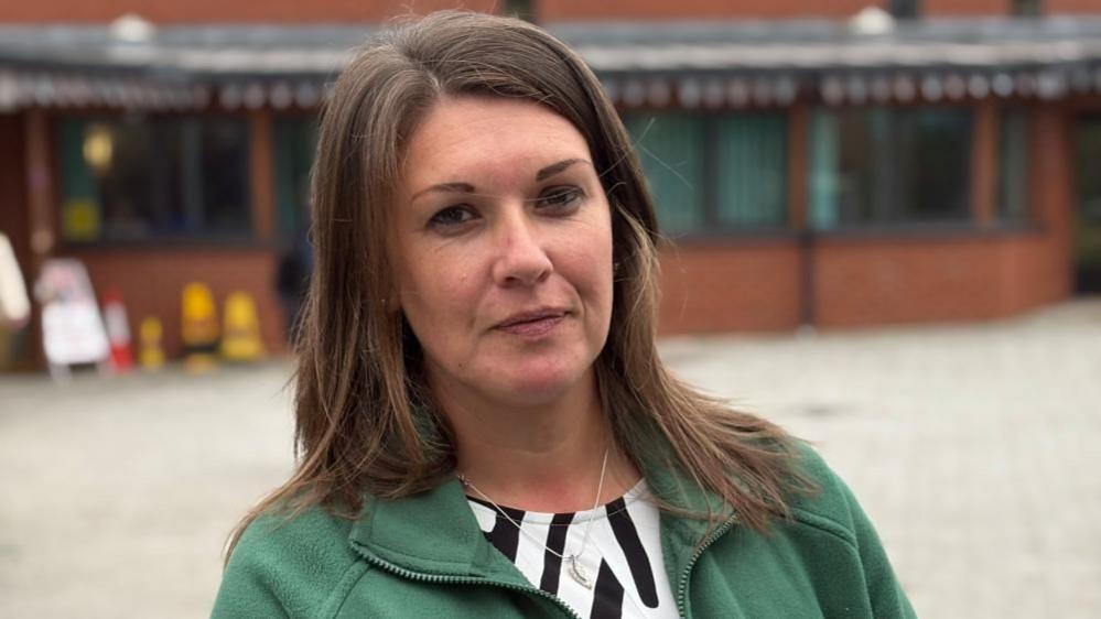 Wearing a green quarter zip jumper and black and white top, Laura Fowler stares at the camera. She has long brown hair which flows down just below her shoulders. The exterior wall of the community centre can be seen in the background.