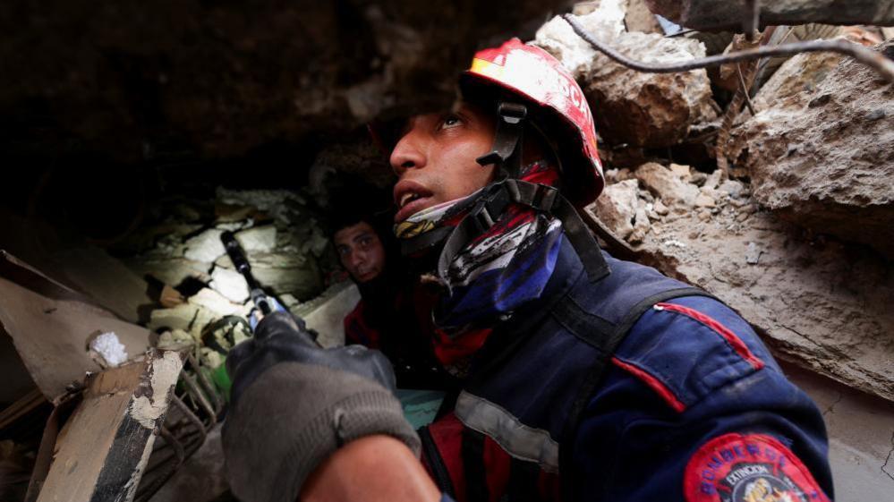 A firefighter wearing a helmet and heavy-duty jacket holds a small camera device up in his hand as he searches for survivors of the earthquakes in La Guaira, Venezuela.