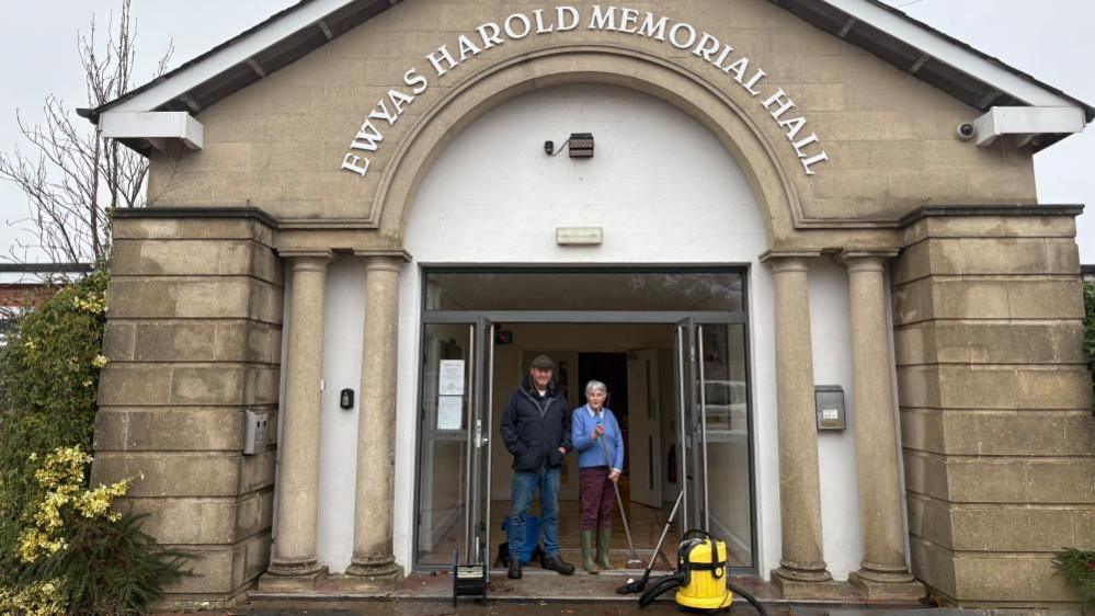 A man and a woman standing outside a memorial hall. The hall is made from stone and has two columns on either side of the entrance. The man and woman are cleaning up after flooding so they are wearing wellies and there is a broom as well as mop bucket and hoover.