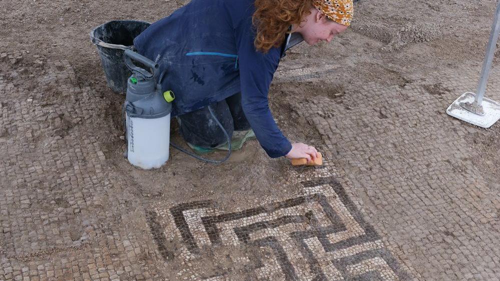 A woman on hands and knees with specialist equipment cleaning a dirty mosaic.