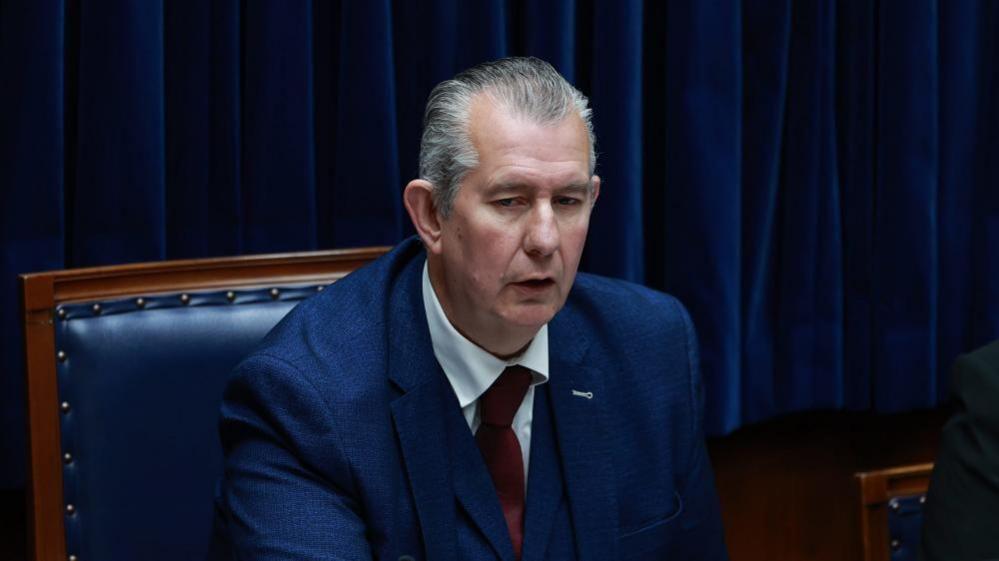 Edwin Poots is sitting in the Speakers chair. It is navy blue with a wooden frame. Poots is wearing a navy blue suit and red tie, with a white shirt. He has combed back grey hair.