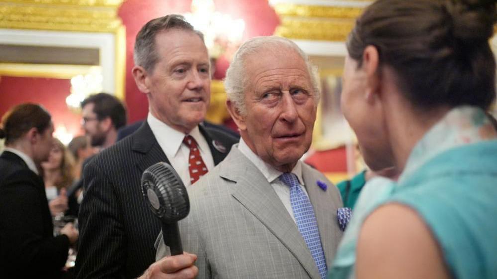 Vice Admiral Sir Tony Johnstone-Burt, Master of the Household to the Sovereign, wearing a black suit and red tie, holds a small battery powered fan for King Charles III, wearing a grey pinstripe suit and blue tie