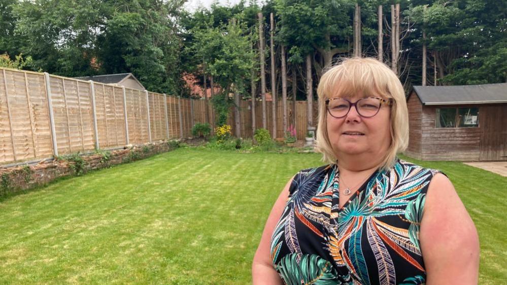 A woman with glasses in a floral dress stands in her back garden 