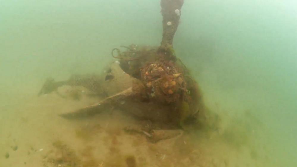 The Hampden P2123 engine and engine cowling is lying underwater on a sandy seabed with three distorted propellers still attached.