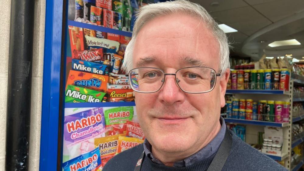 John French  is standing in the entrance of shop you can see branded confectionary  in the background on one side and crisps in the background of the other side. It is a head shot so you can only just see he is wearing a blue shirt with  blue jumper. John is also wearing glasses. 