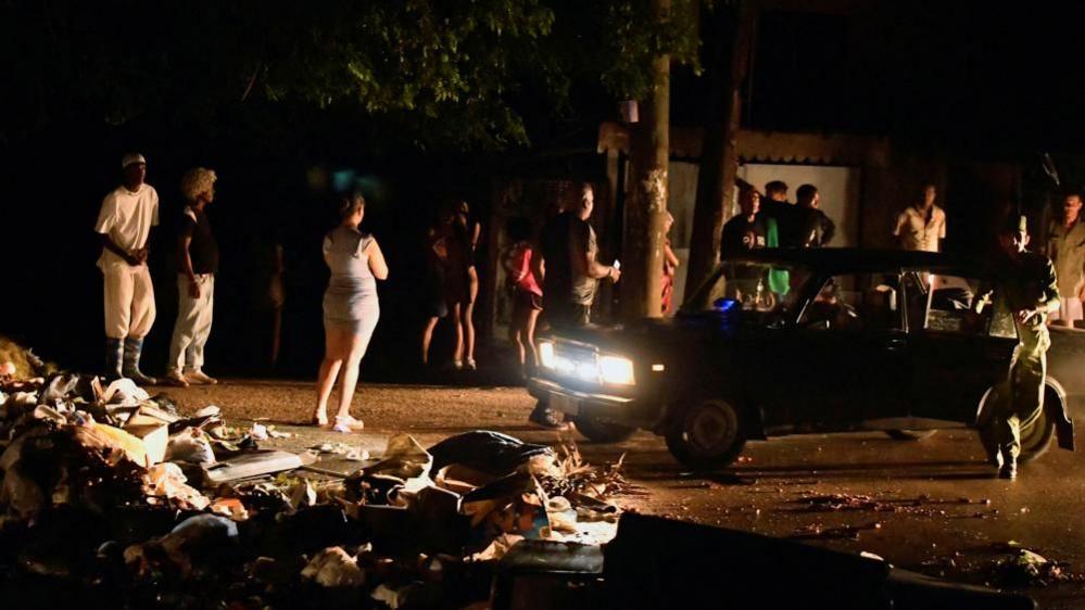 A group of people stand on a dark street in the Cuban capital Havana. Some are illuminated by the headlights of a stationary car. A pile of rubbish and rubble can be seen in the left foreground