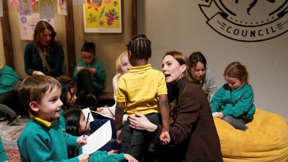 The Princess of Wales has her arm around a little girl, who is wearing a yellow polos shirt with her back to the camera. 