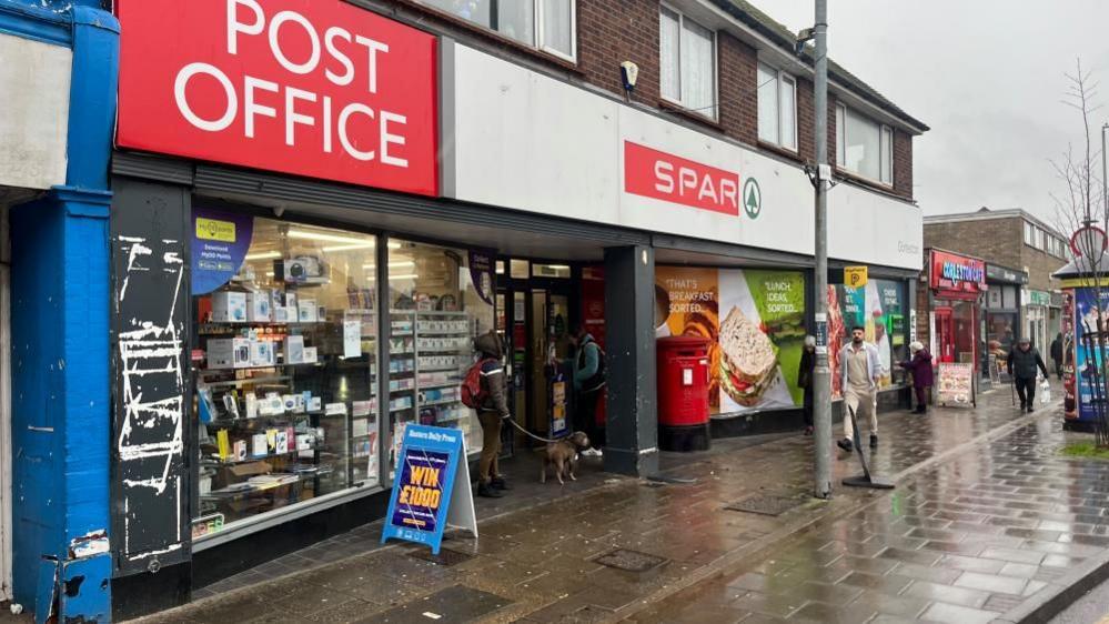 The red signs of the Post Office and Spar, which share the same site. Some windows are stacked with stock and others are covered with vinyl graphic displays. Outside the building is an advertising A-board and a red post box