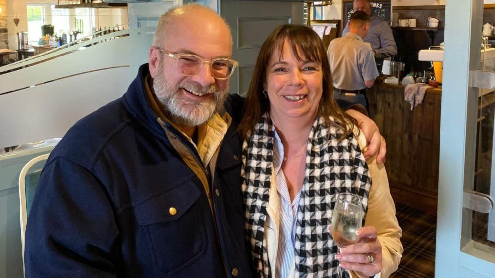 A middle-aged man and woman stand in a bar with their arms around each other, smiling. The woman is holding a glass of wine. 