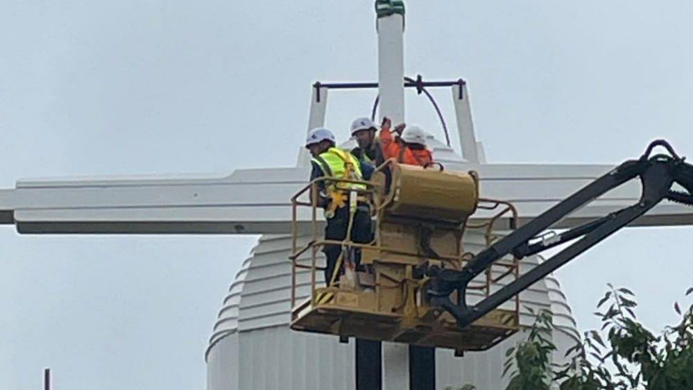 Three men in a cherry picker work on one of the stocks of the Toft Monks Wind Pump, which features a white weather boarded cap.