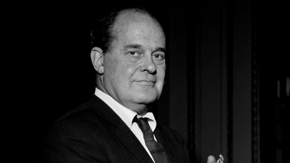 Black and white photo of older man wearing suit and tie looking at the camera .