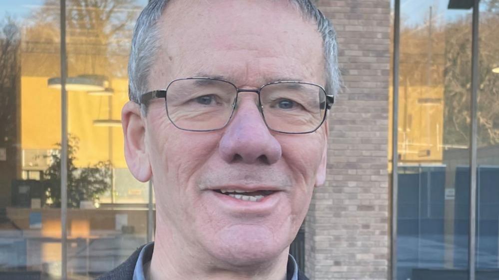 Brian Watkins is wearing glasses and has short greying hair. He wears spectacles, and is smiling. He is stood outside Norfolk County Hall.
